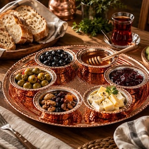 May include: A copper serving tray with small bowls filled with olives, nuts, jam, and cheese. Sliced bread, tomatoes, and cucumbers are also on the table. Two glasses of tea and a honey dipper are visible.