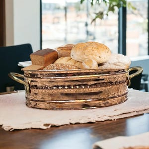Op de afbeelding: Een koperen broodmand met twee handvatten, gevuld met verschillende broden, waaronder een rond brood, een vierkant brood en een paar kleinere broodjes. De mand staat op een wit tafelkleed op een houten tafel.