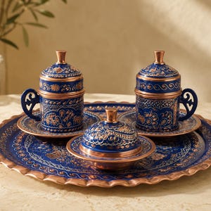 May include: A set of two lidded cups with matching saucers and a lidded bowl arranged on a round tray. Each piece features a deep blue finish with intricate copper-colored floral patterns and metallic copper rims, handles, and finials.