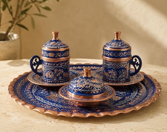 May include: A set of two lidded cups with matching saucers and a lidded bowl arranged on a round tray. Each piece features a deep blue finish with intricate copper-colored floral patterns and metallic copper rims, handles, and finials.