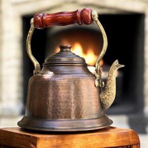 May include: A hammered copper kettle with a wooden handle and a brass spout. The kettle is sitting on a wooden surface in front of a fireplace.