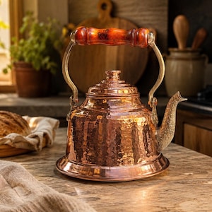 May include: A hammered copper tea kettle with a wooden handle and brass accents. The kettle has a decorative spout and sits on a countertop. The kitchen setting includes a loaf of bread and plants in the background.