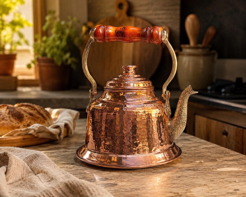 May include: A hammered copper tea kettle with a wooden handle and brass accents. The kettle has a decorative spout and sits on a countertop. The kitchen setting includes a loaf of bread and plants in the background.