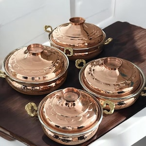 May include: Set of four copper serving dishes with lids and gold handles. The dishes have a hammered finish and a decorative design.
