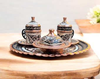 May include: A set of three copper Turkish coffee cups and a sugar bowl with intricate floral designs in black, white, and gold. The cups and bowl are on a copper tray with a scalloped edge.