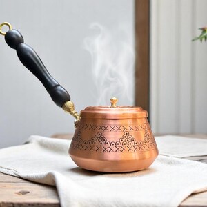 May include: A copper coffee pot with a black wooden handle and intricate engraved designs. Steam rises from the pot, indicating it is hot.