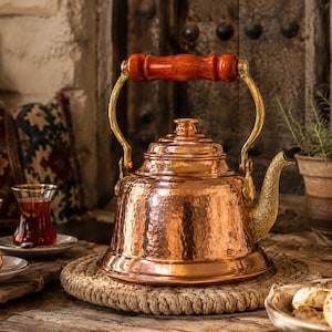 May include: A copper tea kettle with a hammered texture, brass accents, and a wooden handle. The kettle sits on a woven mat, with two glasses of tea and a plate of cookies nearby. A rustic, warm-toned scene.