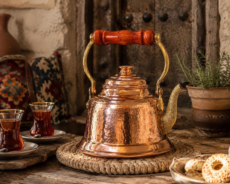 May include: A copper tea kettle with a hammered texture, brass accents, and a wooden handle. The kettle sits on a woven mat, with two glasses of tea and a plate of cookies nearby. A rustic, warm-toned scene.