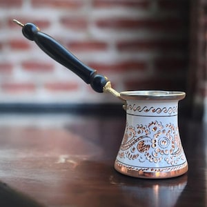 以下が含まれることがあります： 黒の木製のハンドルが付いた白と金のトルコ式コーヒーポット。ポットには花柄と銅の縁取りがあります。