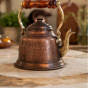 May include: A copper tea kettle with a hammered texture, brass accents, and a wooden handle. The spout features an ornate design. The kettle sits on a marble surface. A decorative kitchen item.