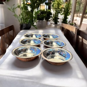 May include: Six silver-coloured bowls with a hammered metal texture and copper rims are arranged on a white tablecloth. The bowls are arranged in two rows of three.