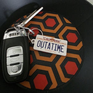 Pode incluir: Uma chave de carro Audi e um chaveiro com um design de placa de carro da Califórnia que diz "OUTATIME" em letras brancas. O chaveiro está ao lado de um porta-copos com um padrão geométrico preto e laranja.