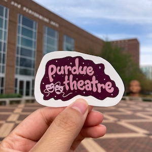 May include: A close-up of a sticker with the text "Purdue theatre" in pink lettering on a dark purple background. The sticker has a white border and is decorated with two pink theatrical masks and white stars.