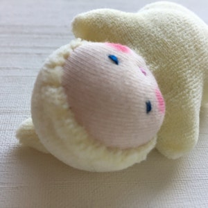 May include: A soft, plush lamb toy in cream and pink. The lamb's face is a light pink with embroidered blue eyes and a pink nose. The body is cream-colored and textured.