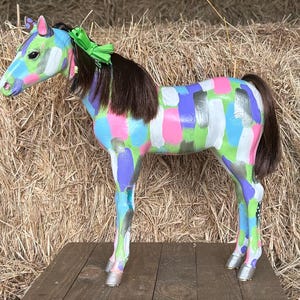 以下が含まれることがあります： 鮮やかな色でペイントされた馬の置物。馬は、緑、ピンク、青、紫、銀などの明るい色の混合で装飾されています。濃い茶色のたてがみと尾、緑のリボンが付いています。馬は木の表面に立っています。