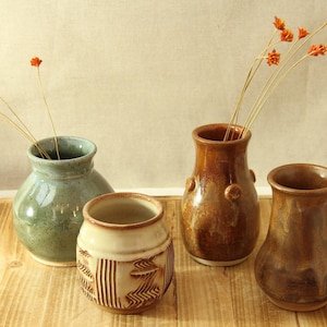May include: Four ceramic vases of varying shapes and colors are arranged on a wooden surface. Two vases hold dried orange flowers. The vases are glazed in shades of green, brown, and cream, showcasing a rustic aesthetic.