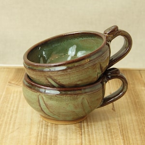 May include: Two handmade ceramic mugs stacked on a wooden surface. The mugs are a muted green with brown rims and handles. The mugs have a rustic, organic feel, with visible brushstrokes and a textured surface.