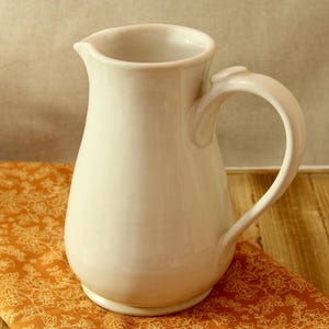 May include: A cream-colored ceramic pitcher with a curved handle and a spout. The pitcher sits on a patterned orange cloth, with a wooden surface in the background. The pitcher is a kitchenware item.