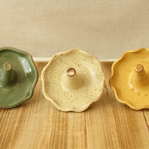 May include: Three ceramic incense holders in green, cream, and yellow. Each has a scalloped edge and a central post for holding incense. The yellow holder features a bee design. The holders are displayed on a wooden surface.