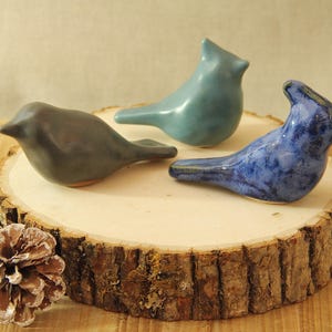May include: Three ceramic bird figurines in shades of blue and gray rest on a wooden slice. The birds have smooth, rounded bodies and are arranged in a decorative display. A pine cone is in the foreground.