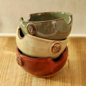 May include: Stacked ceramic bowls in shades of green, cream, and red. Each bowl has a unique leaf design and a curved rim. The bowls are handmade and sit on a wooden surface.