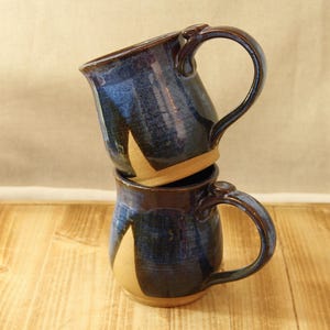 May include: Two handmade ceramic mugs stacked on top of each other. The mugs are glazed in a dark blue color with lighter blue and tan accents. Each mug has a large handle and a rounded shape, perfect for coffee or tea.