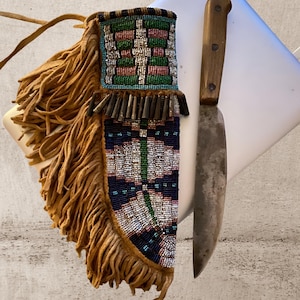 Peut inclure: Un couteau à manche en bois et lame argentée usée est posé à côté d'un étui en cuir à franges. L'étui est orné de perles colorées bleues, vertes, rouges et blanches, et possède une frange beige.