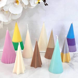 May include: A collection of cone-shaped candles in various colors, including pink, yellow, green, white, brown, blue, and gray. The candles have a textured, ribbed design and are arranged on a white surface.