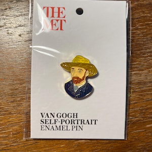 May include: Enamel pin featuring Van Gogh's self-portrait, packaged on a white card with "THE MET" logo. The pin depicts the artist in a yellow hat and blue jacket. The text "VAN GOGH SELF-PORTRAIT ENAMEL PIN" is printed below the pin.