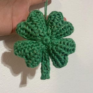 Può includere: Un quadrifoglio verde all'uncinetto con accenti di filo metallico dorato. Il quadrifoglio è sospeso da un filo verde, suggerendo che sia un ornamento. L'oggetto fatto a mano ha un aspetto strutturato.