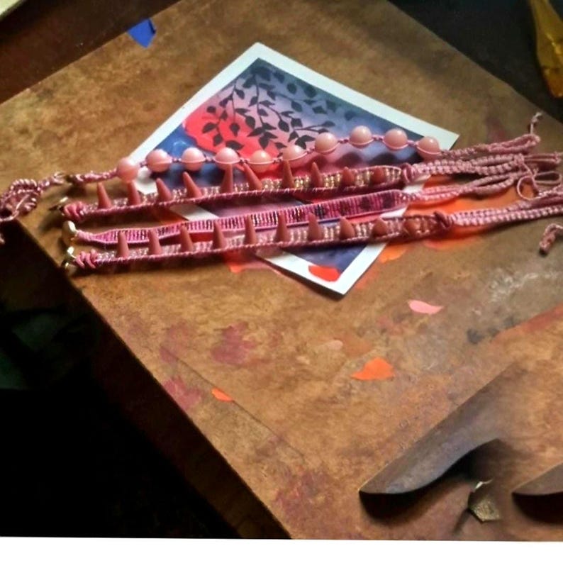 May include: Three pink bracelets with a woven design and small, cone-shaped embellishments. One bracelet features large, round beads. The bracelets are displayed on a wooden surface with a watercolor painting in the background.