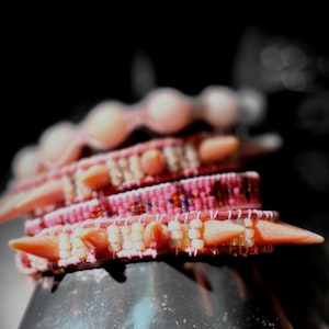 May include: Close-up of several stacked pink bracelets with pointed, coral-colored spikes. The bracelets are intricately woven with white and pink beads, creating a textured and detailed appearance. The background is blurred, emphasizing the bracelets.
