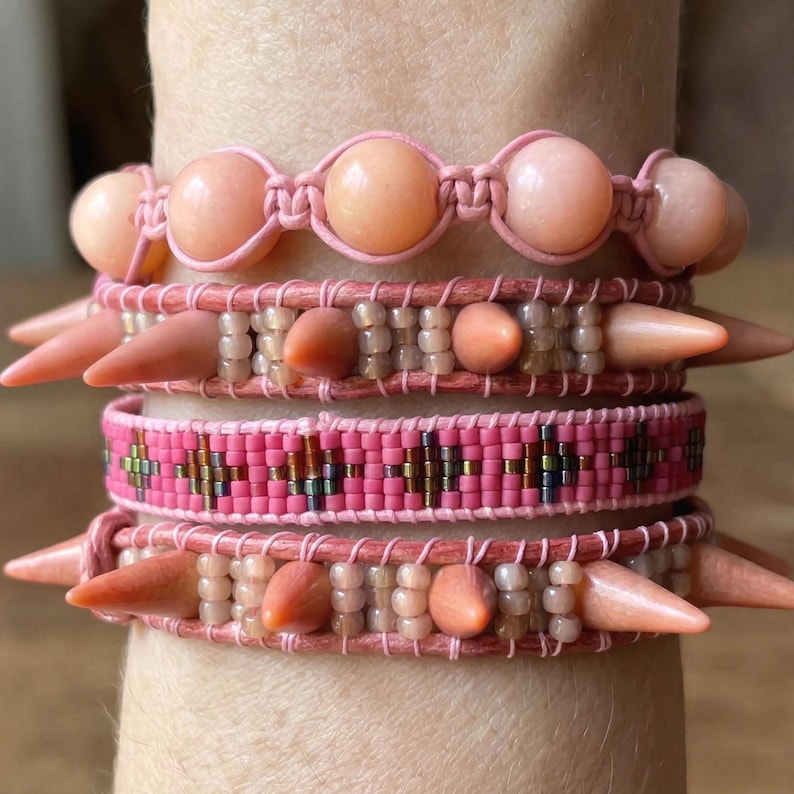 May include: A collection of pink bracelets with various designs. One bracelet features large, round beads, while others have spike accents and beaded patterns. The color palette includes shades of pink and peach, creating a cohesive and stylish accessory set.