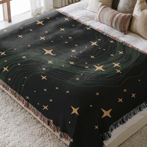 May include: A dark green and black throw blanket with a celestial design. The blanket features a pattern of gold stars and flowing green lines. The edges are finished with a fringe. The blanket is displayed on a bed with pillows.