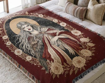 May include: A woven throw blanket featuring a dark red background with a central illustration of a winged figure with long hair, surrounded by a circular frame of cream-colored roses and a golden halo against a starry night sky backdrop.