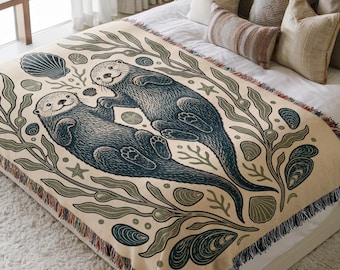 May include: A cream-coloured woven throw blanket featuring a dark blue illustration of two sea otters floating together. The design is surrounded by green seaweed, various seashells, and small starfish, finished with a fringed edge on the sides.