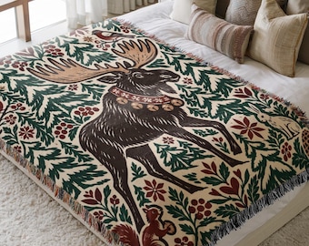 May include: A woven throw blanket featuring a large central moose illustration surrounded by a repeating pattern of green pine branches, red berries, small birds, a squirrel, and a rabbit on a cream-coloured background with fringed edges.