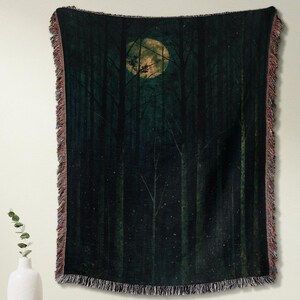 May include: A dark green woven throw blanket featuring a forest scene with a full moon. The blanket has a fringed edge with a reddish-brown color. The throw is hanging on a wall above a wooden cabinet.