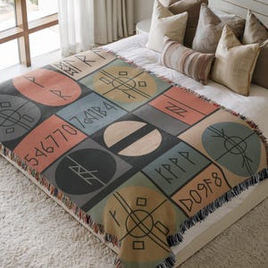 May include: A colorful throw blanket with a geometric design featuring squares and circles in shades of gray, peach, blue, and tan. The blanket has a fringed edge and is decorated with symbols and numbers.