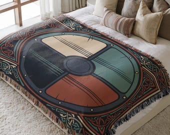 May include: A woven throw blanket featuring a large circular shield design divided into four quadrants of cream, dark blue, teal, and terracotta. The shield has a central dark gray boss and is surrounded by intricate dark brown Celtic knotwork.