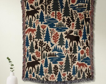May include: A woven throw blanket featuring a repeating pattern of dark brown moose, orange foxes, and black owls amidst blue and green pine trees, red flowers, and winding rivers on a cream background, finished with a fringed edge on all sides.