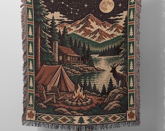 May include: A woven throw blanket featuring a nighttime mountain landscape with a cabin, tent, campfire, and deer. The scene includes a full moon, starry sky, and pine trees. The border has a repeating pine tree and geometric pattern with fringe.