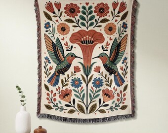 May include: A woven throw blanket featuring a symmetrical design of two hummingbirds facing a central large flower, surrounded by smaller floral and leaf motifs in shades of red, blue, green, and cream, finished with a fringed edge on all sides.