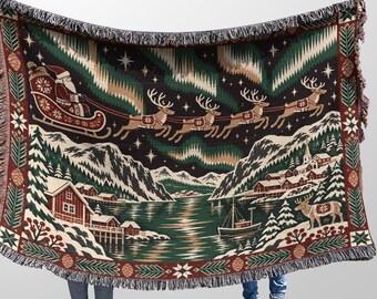 May include: A woven throw blanket featuring a festive winter scene with Santa in a sleigh pulled by reindeer across a night sky with green aurora borealis. The landscape includes snow-covered mountains, a village, a boat, and pine tree borders.