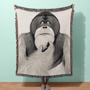 May include: A woven throw blanket featuring a black and white illustration of an orangutan with its arms wrapped around itself. The orangutan has a white beard and is looking up.