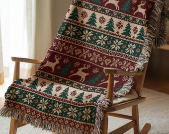May include: A woven throw blanket featuring a repeating pattern of reindeer, pine trees, and snowflakes in shades of deep red, forest green, and cream. The blanket has a textured knit appearance with fringed edges along the bottom and sides.