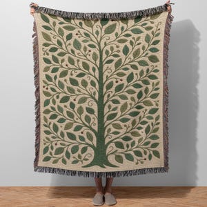 May include: A beige throw blanket featuring a stylized tree design in shades of green. The tree has a thick trunk and many leaves, with small red accents. The blanket has a fringed edge.