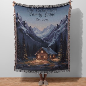 May include: A woven throw blanket featuring a mountain landscape with a cabin, trees, and text that reads "The Richardson Family Lodge Est. 2025." The blanket has a dark blue background with a fringed edge.