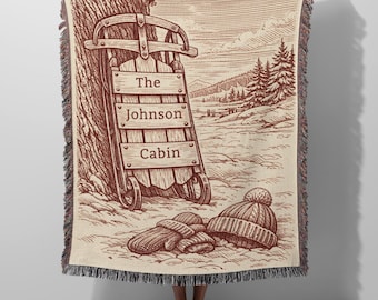 May include: A woven throw blanket featuring a sepia-toned illustration of a wooden sled leaning against a tree in a snowy landscape. The sled displays the text The Johnson Cabin. A knit hat and a pair of mittens rest on the snow in the foreground.