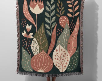 Puede incluir: Una manta tejida de color verde oscuro con un diseño botánico. El diseño incluye hojas, flores y vainas de semillas estilizadas en tonos crema, melocotón y verde oliva. La manta tiene un borde con flecos.